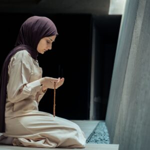 A woman in hijab kneels in prayer with beads inside a peaceful mosque environment.