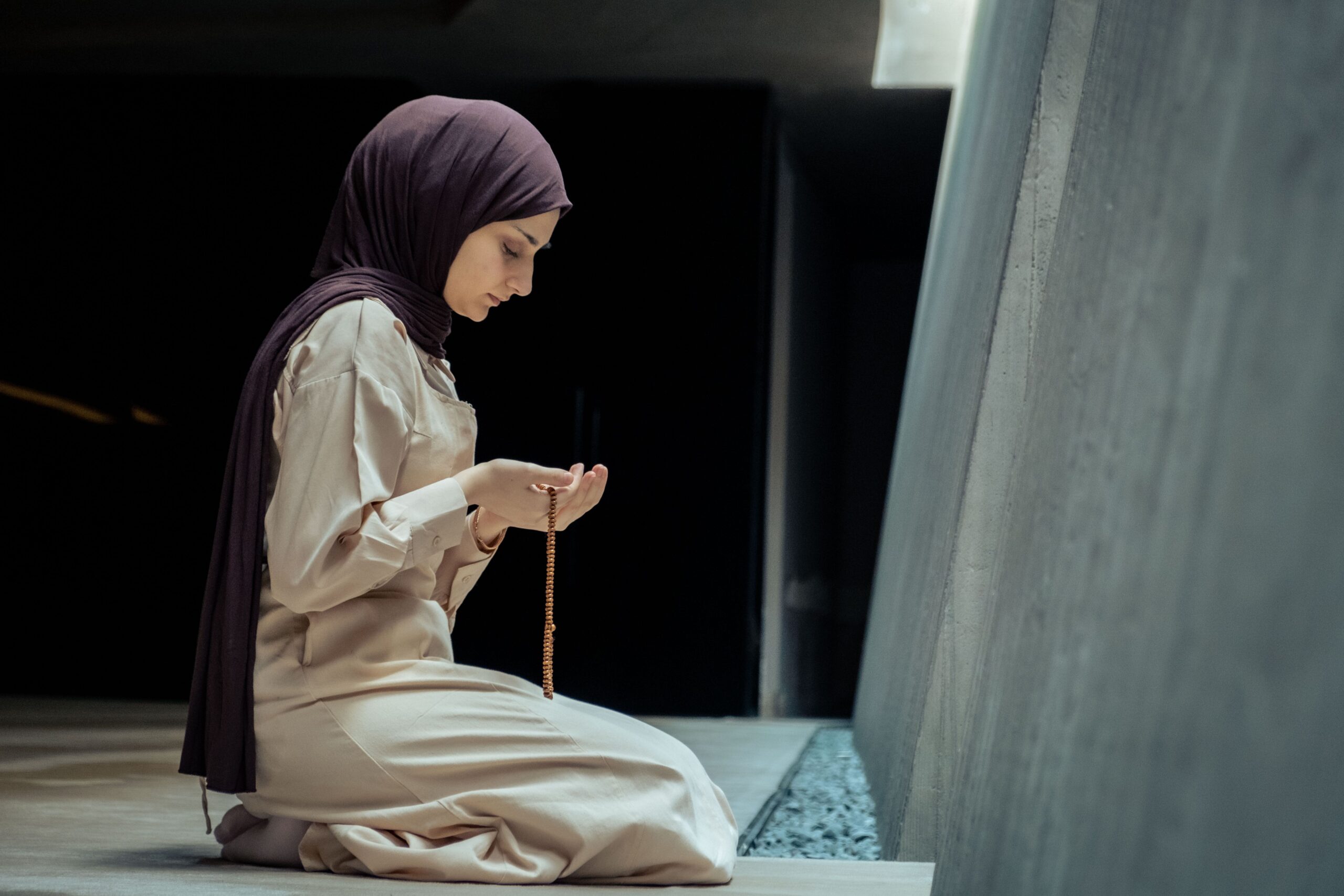A woman in hijab kneels in prayer with beads inside a peaceful mosque environment.