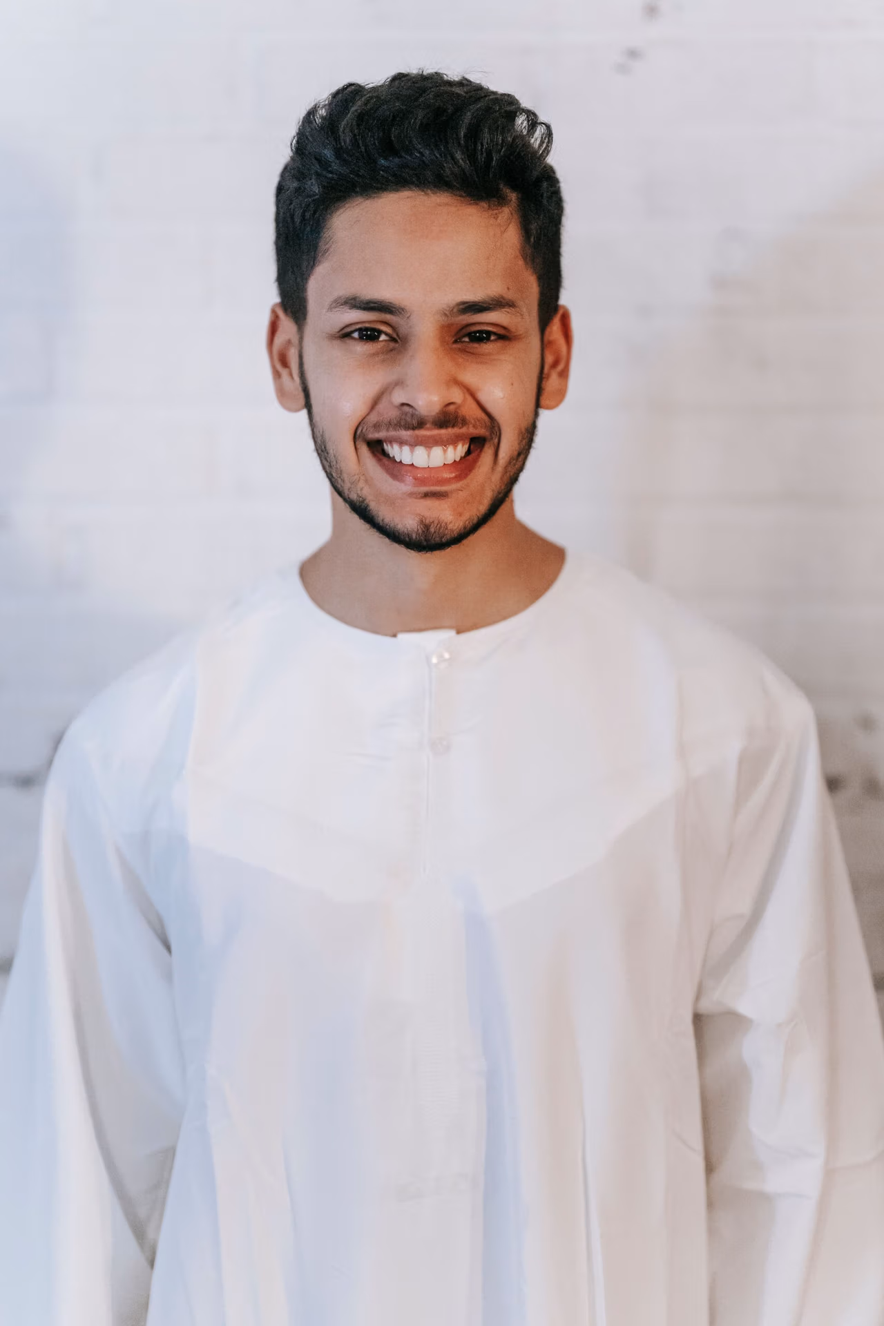 Portrait of a smiling man in a traditional white thobe against a white brick wall indoors.